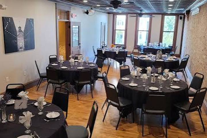 Round tables dressed in black linen and set with glassware beneath a pressed-tin ceiling and tall windows