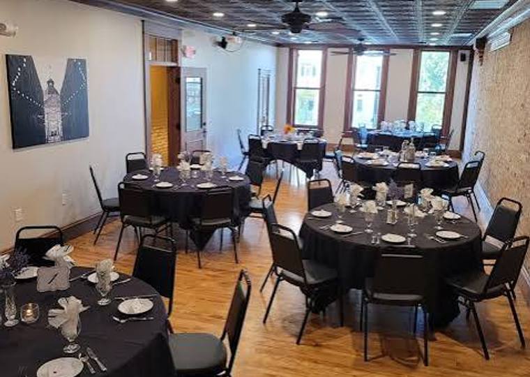 Round tables dressed in black linen and set with glassware beneath a pressed-tin ceiling and tall windows