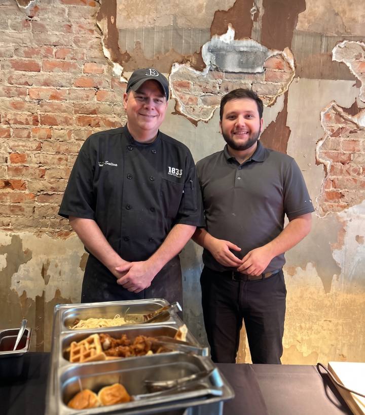 Two of the house team, one in a chef's coat and one in a house polo, standing behind the chafing dishes of the Sunday brunch buffet in front of the exposed brick wall