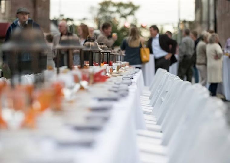 A long table set down the middle of the street at dusk, lanterns lit along it and guests gathered at either side